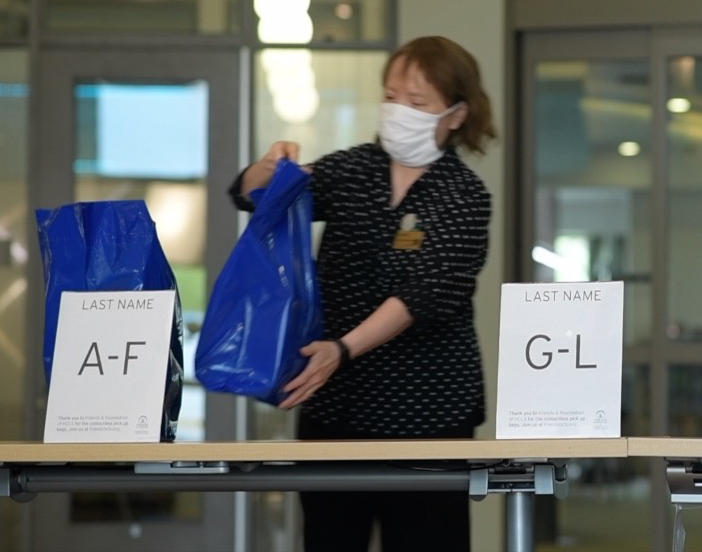 A library staff member, wearing a face covering, places blue bags on a table with alphabetic signs, A-F and G-L.

