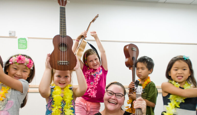 Instructor smiling and holding up a ukulele from a free Ukulele kit apart of the Library of Things collection with her students completing the same action behind her.