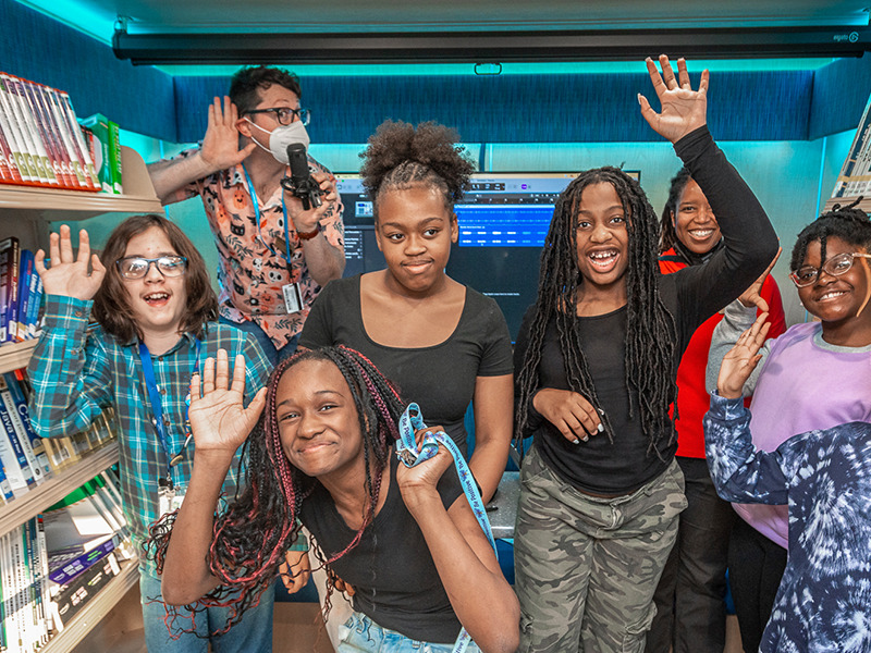 group of students in the STEAM machine smiling and posing with their hands up while two instructors are completing the same action behind them.