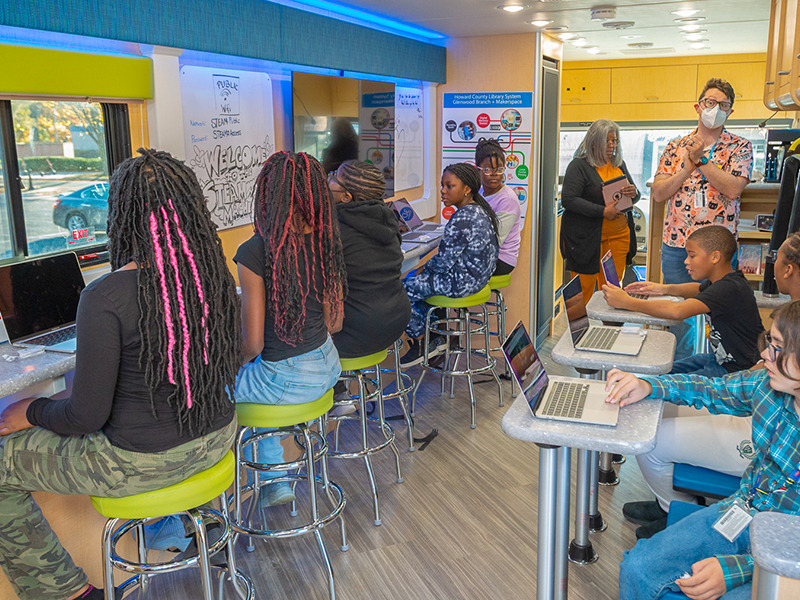 children sitting in the STEAM machine with free laptops in front of them, while a Howard County Library System teacher is instructing them on a STEM based activity.