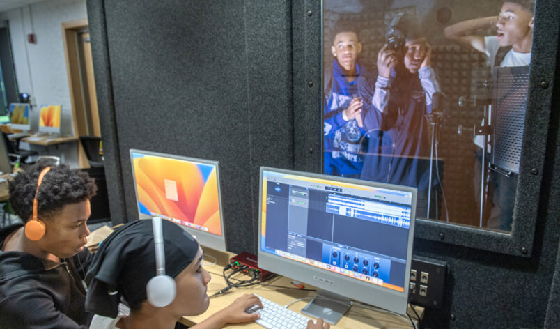 Two teens working computers In front of a sound booth during a recording session