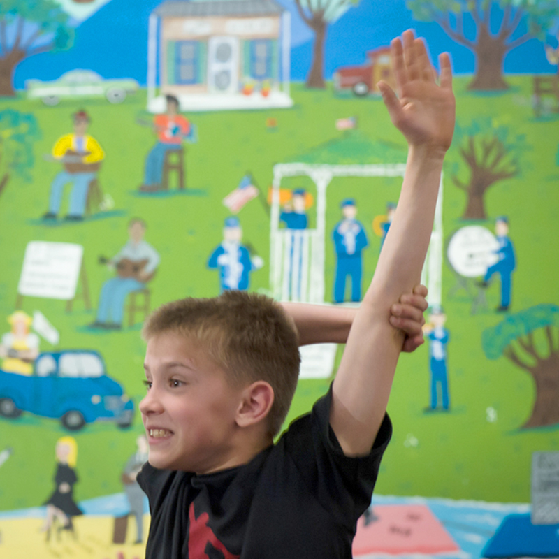 Child raising his had with an excited smile, in front of a community mural.