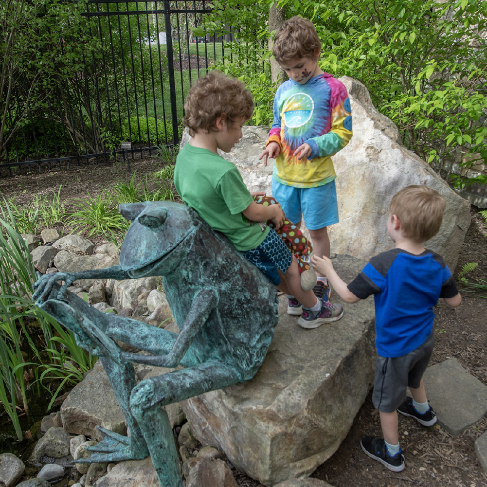 Children standing on rocks and leaning on frog statue.