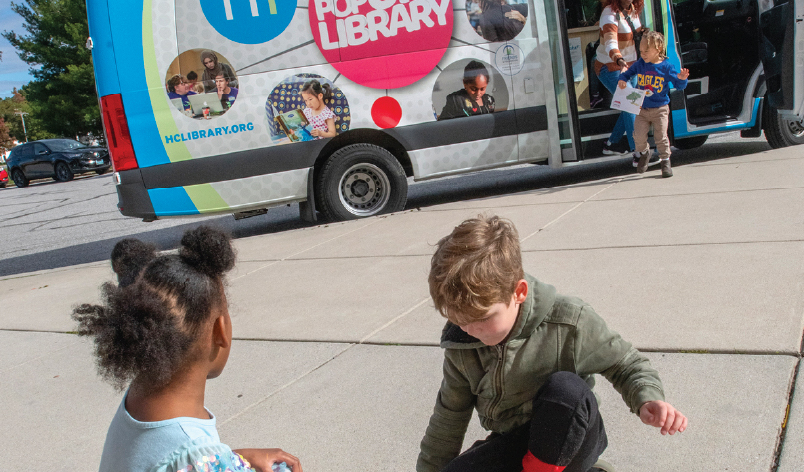 Young children engaging in activities outside the Howard County Library System Pop-Up Library during a Library on the Go outreach stop, bringing books and learning resources into the community.