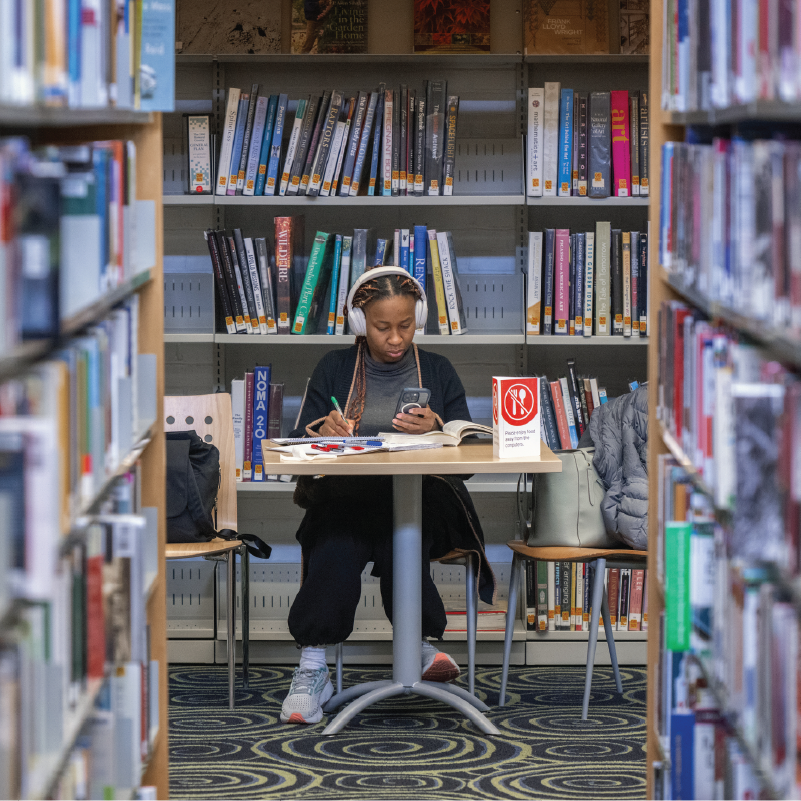 A student wearing earphones at a table covered in books.